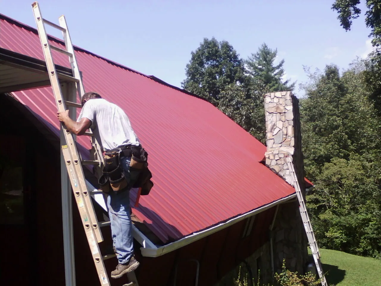 Roofer on a ladder beside a red metal roof and stone chimney — Soffit & Fascia Repair by Roofing Brothers in Pompton Lakes